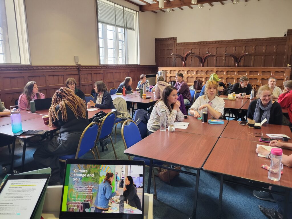 A room full of people sit around tables talking. A laptop screen says "climate change and you..."